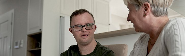 Ein junger Mann mit einer sichtbaren Behinderung im Gespräch mit einer älteren Frau.